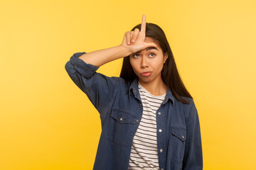 I lost! Portrait of unlucky sad girl in denim shirt standing with loser gesture, L sign on forehead and blaming herself for failure, fired from work. indoor studio shot isolated on yellow background