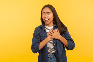 Myocardial infarction. Portrait of girl in denim shirt pressing on chest with painful expression, suffering sudden ache, heart attack, cardiac problems. studio shot isolated on yellow background