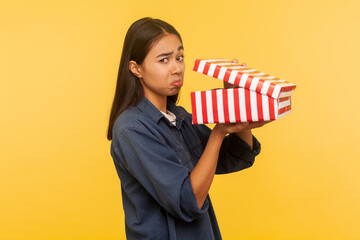 Portrait of upset girl in denim shirt opening gift and looking at camera with unhappy disappointed...