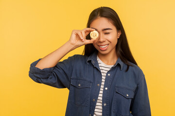 Cryptocurrency btc coin. Portrait of positive rich girl in denim shirt smiling and looking through golden bitcoin, electronic money, digital currency. indoor studio shot isolated on yellow background