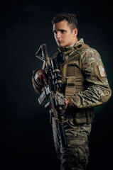 a brutal guy in military airsoft overalls poses with a weapon in his hands on a dark background