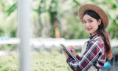 Fototapeta premium Portrait of young beautiful woman holding clipboard, smiling and looking at camera while standing at greenhouse hydroponic organic farm.