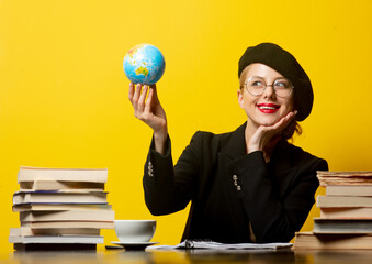 Style blonde woman in beret sitting at table with books around and holds globe on yellow background