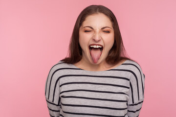 Portrait of disobedient funny woman in striped sweatshirt showing tongue out, expressing displeasure, teasing with derisive goofy grimace, naughty behavior. studio shot isolated on pink background