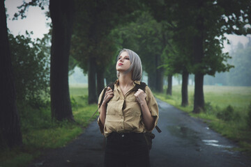 Fototapeta na wymiar Style blonde woman with backpack at countryside road in rainy day