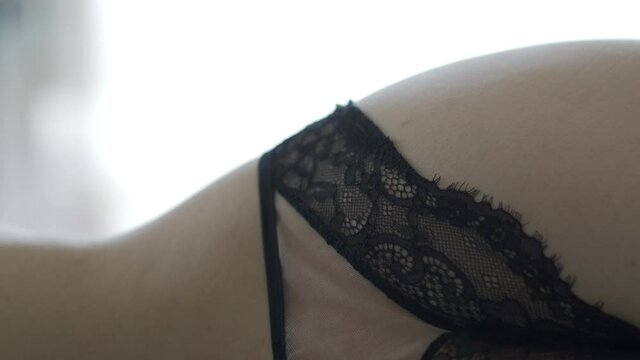 Rear view of beautiful sexy woman in black lingerie lying on her side on bed. Close up of female bare back against window in the morning. Adult female model wearing sensual lace panties.