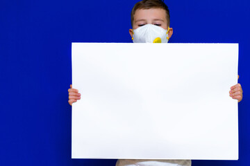 Close up caucasian boy in a protective mask holds a blank sheet of paper on a bright blue isolated background.