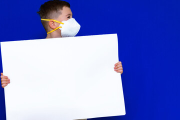 Close up caucasian boy in a protective mask holds a blank sheet of paper on a bright blue isolated...