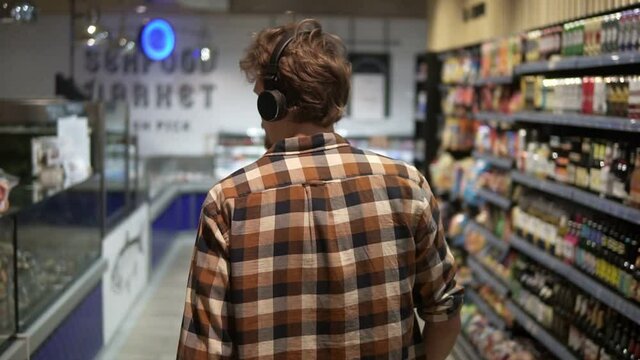 Stylish caucasian guy with headphones walks through goods section of the store, wearing plaid shirt - listening to the good music.. Following back view shot. Slow Motion