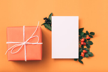 Holidays greeting. White blank paper. Gift box. Rose hip decor. Orange background.