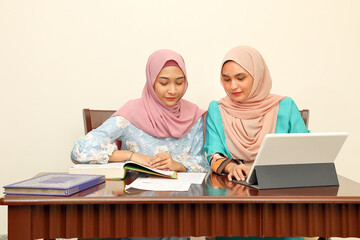 Two young Asian Malay Muslim woman wearing headscarf at home office student sitting at table talk mingle look at computer book document study discuss smile happy