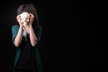 beautiful young curly girl on a black background holds a skull in her hands and hides behind him