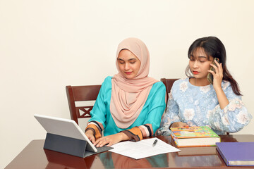 Two young Asian Malay Muslim woman wearing headscarf at home office student sitting at table phone computer book document study talk work mingle