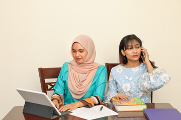 Two young Asian Malay Muslim woman wearing headscarf at home office student sitting at table phone computer book document study talk work mingle