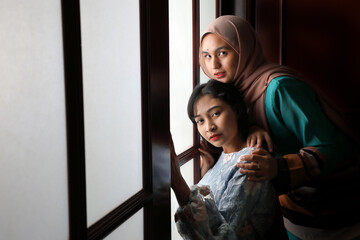 two young Asian Malay muslim woman wearing headscarf baju kurung dress at home in front window mood light on face dark background peaceful content calm tranquil break trough wonder happy