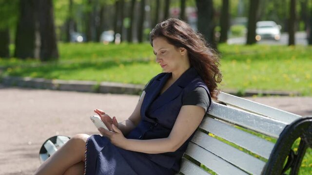 Adult happy woman using smartphone sitting on bench in park woman texting sharing messages on social media brunette woman holding mobile phone smiling.