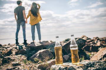close-up two bottles of beer stand on stones near the water in the sun on a background of a couple. man and woman hold hands