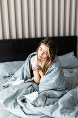 Sick woman froze and caught a cold from an unheated flat covered with warm blanket sit in bed drinking cup with hot beverage or medicines using handkerchief blows runny nose. Seasonal grippe concept