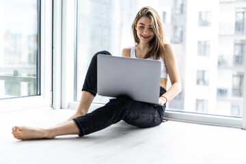 Beautiful young woman in white shirt, jeans sitting on windowsill with laptop