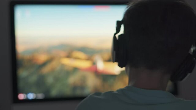 Boy plays game of War Thunder strategy online on computer with headphones in the evening in room. Light of monitor and silhouette of child play emotionally at computer