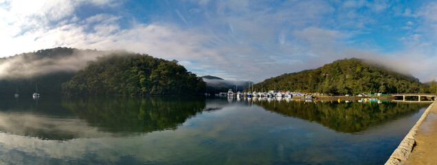 Beautiful morning panoramic view of Cockle creek with reflections of blue sky, light clouds, foggy mountains and trees, Bobbin Head, Ku-ring-gai Chase National Park, New South Wales, Australia