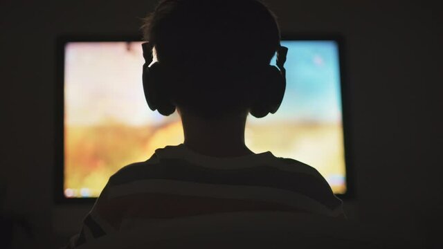Boy plays game of War Thunder strategy online on computer with headphones in the evening in room. Light of monitor and silhouette of child play emotionally at computer