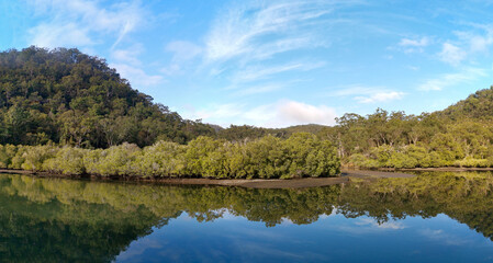 Fototapeta premium Beautiful morning view of Cockle creek with reflections of blue sky, foggy mountains and trees, Bobbin Head, Ku-ring-gai Chase National Park, New South Wales, Australia