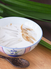 Sweet rice noodles with coconut cream (Pla Krim Khai Tao) in a ceramic bowl with a wooden spoon on cutting board and pandanus leaves are the backdrop...