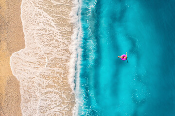Aerial view of a young woman swimming with the donut swim ring in transparent blue sea with waves at sunset in summer in Turkey. Tropical landscape with girl, azure water, sandy beach. Top view