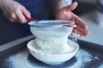 A man sifts crumbly wheat flour through a metal sieve into a white bowl. Cooking process.