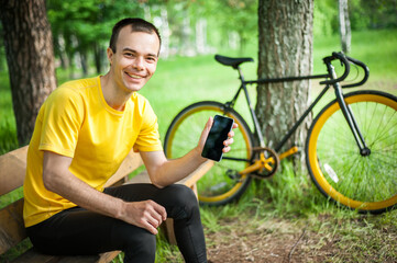 A young man sitting on a bench in a public Park communicates via mobile communication. In the background with his bike. Among trees and vegetation.