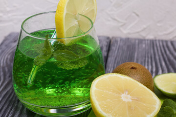 Green lemonade in a glass. Mint leaves float in it. Garnished with a slice of lemon. With a straw for a cocktail. Nearby on a cutting board are fruits for a cocktail.