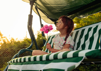 Little girl with pink peony is resting on a bench in garden.