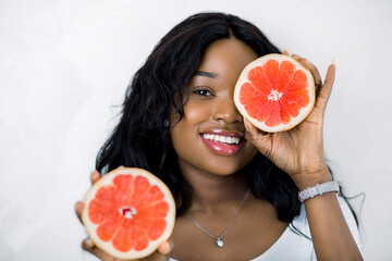 Close-up portrait of charming african girl holding halfs of grapefruit, hiding her eye. Amazing smiling dark skinned woman with grapefruit slices looking at camera on white background