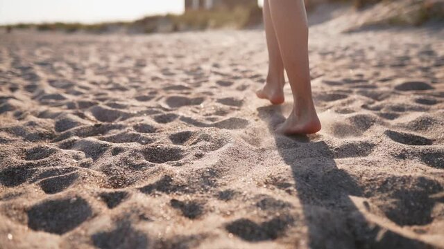Back view of tanned woman legs and feet walking on sandy beach to the sea on sunny day. Slim pretty girl goes by the sea on clear white sand. Female on vacation. Low camera angle, slow motion.