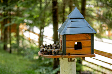 Wooden bird feeder in the form of a house with a roof, attached to a wooden post in a forest among trees in the nature