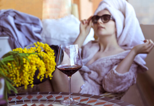 Woman with a glass and flowers. Girl smokes aykos in a bathrobe and with a towel on her head, drinks wine on the veranda. The state of relaxation and sexual mood. Creative photo, soft focus.