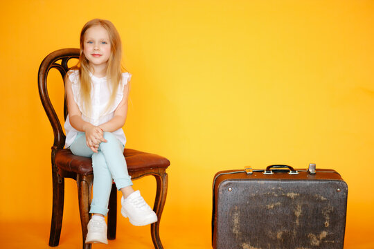 Little young blonde Girl sitting on old vintage chair in yellow studio and smiling next to old vintage suitcase travelling trunk . Travaller style concept. Voyager adventure lifestyle, Travel style
