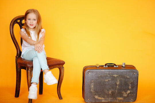 Little young blonde Girl sitting on old vintage chair in yellow studio and smiling next to old vintage suitcase travelling trunk . Travaller style concept. Voyager adventure lifestyle, Travel style