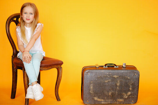 Little young blonde Girl sitting on old vintage chair in yellow studio and smiling next to old vintage suitcase travelling trunk . Travaller style concept. Voyager adventure lifestyle, Travel style