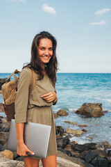 Freelancer girl while traveling to the sea with a backpack and laptop for work, smiling while standing on the sea pier at sunset