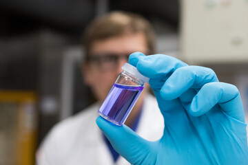 Scientist with gloved hands in a laboratory holding a small vial containing a purple chemical