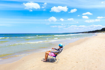 Fototapeta na wymiar Young woman tourist sitting in beach chair on beautiful white sand beach with blue sea near Kolobrzeg, Baltic Sea coast, Poland