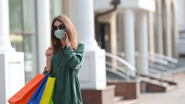Cute girl in a dress and a protective mask on her face holds shopping bags and communicates on the phone.