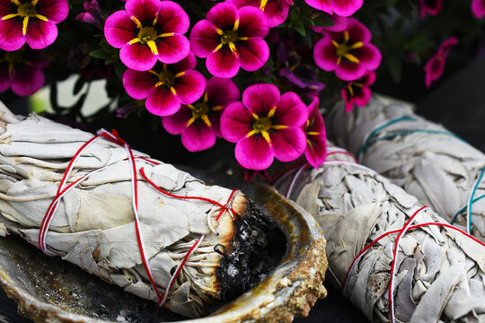 A close up image of three white sage bundles and abalone shell next to bright pink flowers. 