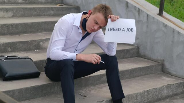 Unemployed sad job seeker with the inscription "I need a job" smokes while sitting on the steps