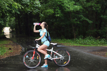 Fototapeta premium sport girl in sportswear stands with her back beside a bicycle drinks water from a bottle