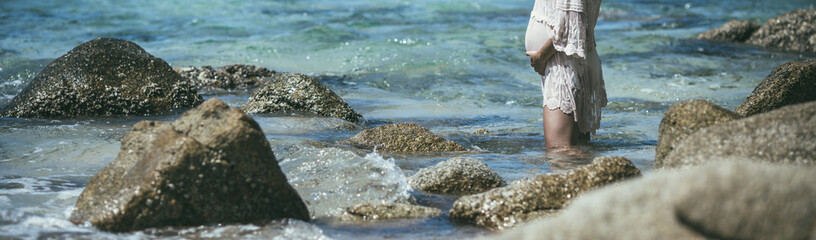 Panorama of the sea with a pregnant girl standing in the water. Woman holds a pregnant belly and enjoys the spray of the ocean. Sea holiday. Image with selective focus, noise effect and toning