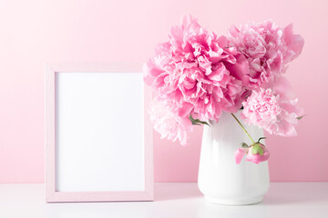 Home decoration with floral decor, frame poster on table. Beautiful flowers pink peonies in vase on pink background