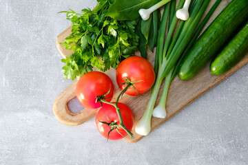 Wooden cutting board with fresh vegetables on stone concrete table. Top view tomatoes, cucumbers, onion, parsley. Fresh healthy food concept.